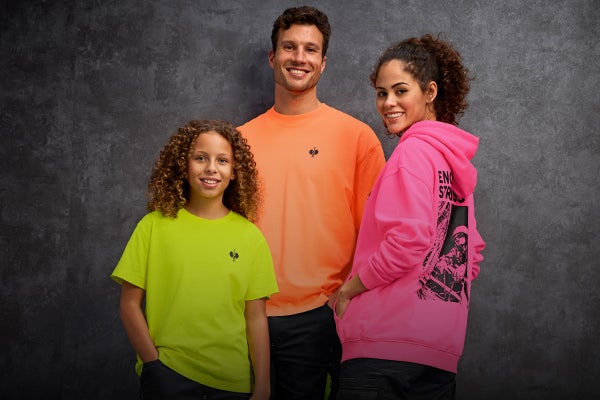 Ein Mann in einem neonorangen Vintage-Shirt, eine Frau in einem neonpinken Vintage-Hoodie und ein Kind in einem neongelben Vintage-Shirt stehen gemeinsam vor einer grauen Betonwand.