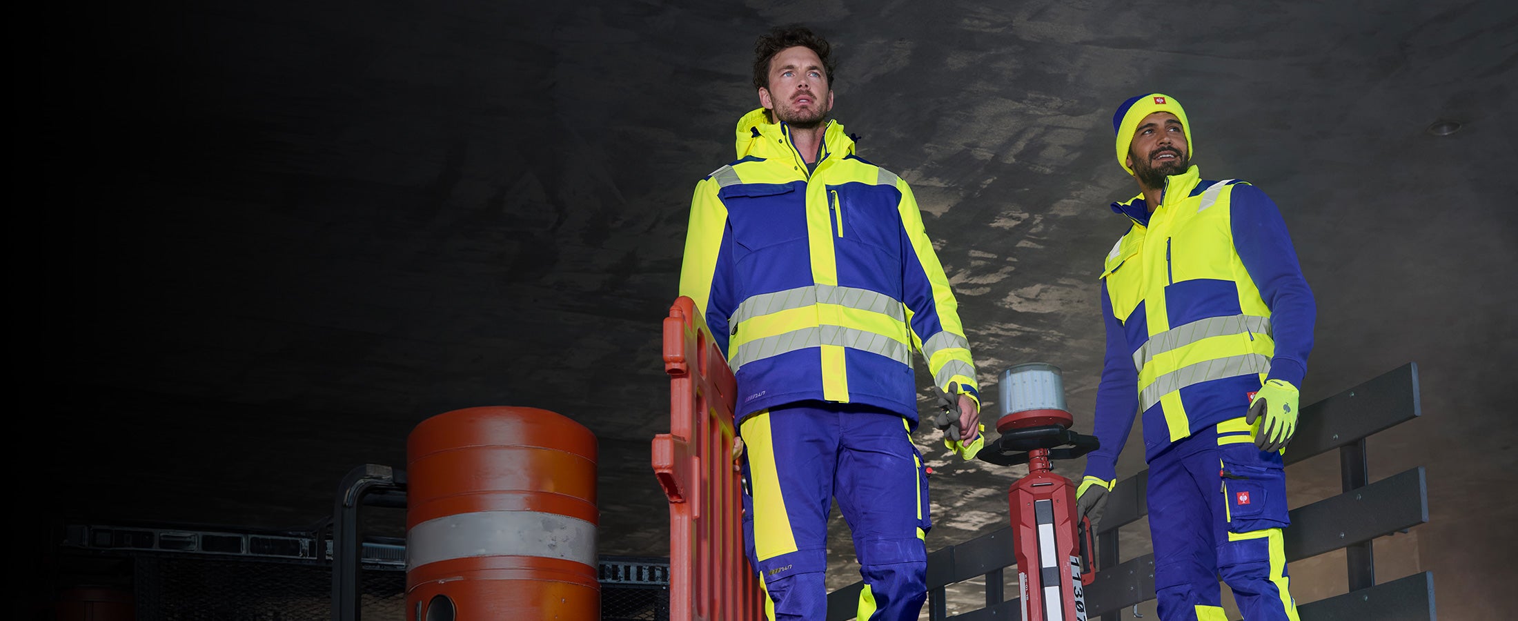Zwei Männer in Warnschutzkleidung aus der motion24/7 Kollektion vor dunklem Bauwerk, einer davon mit Mütze.
