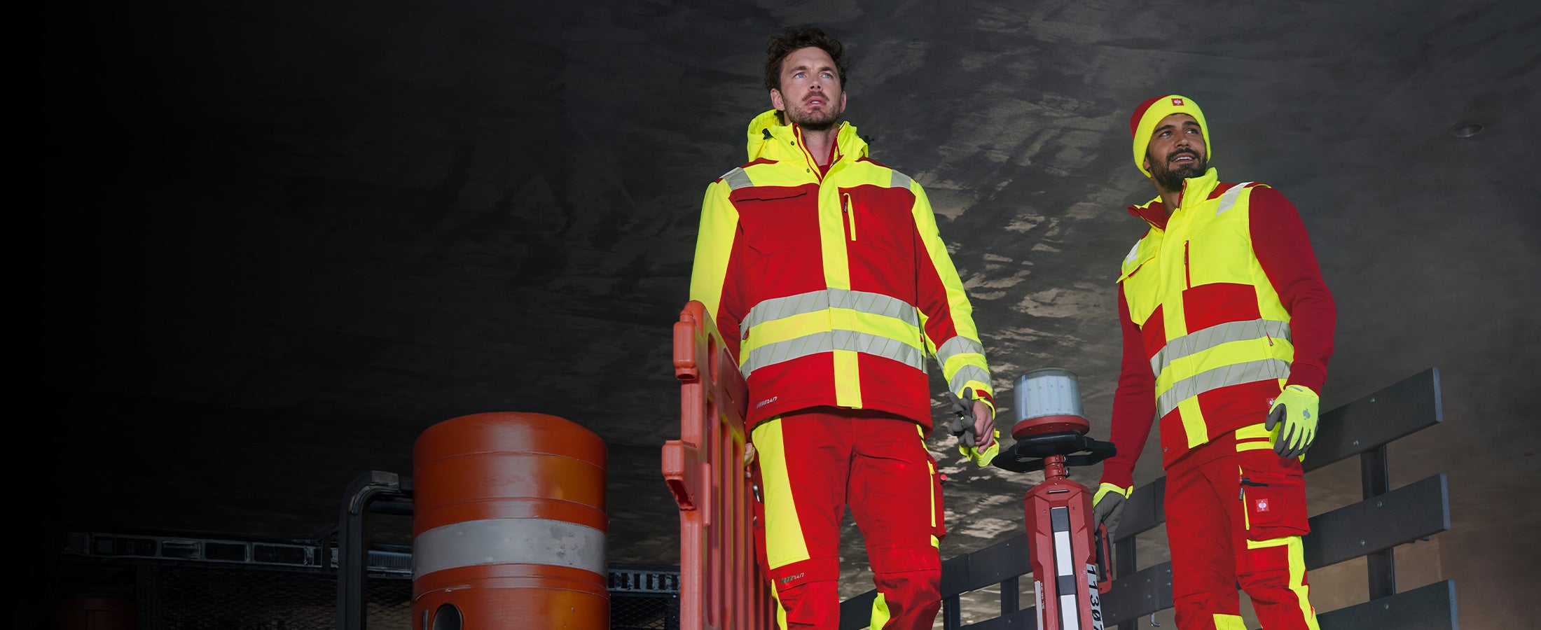Zwei Männer in Warnschutzkleidung aus der motion24/7 Kollektion vor dunklem Bauwerk, einer davon mit Mütze.