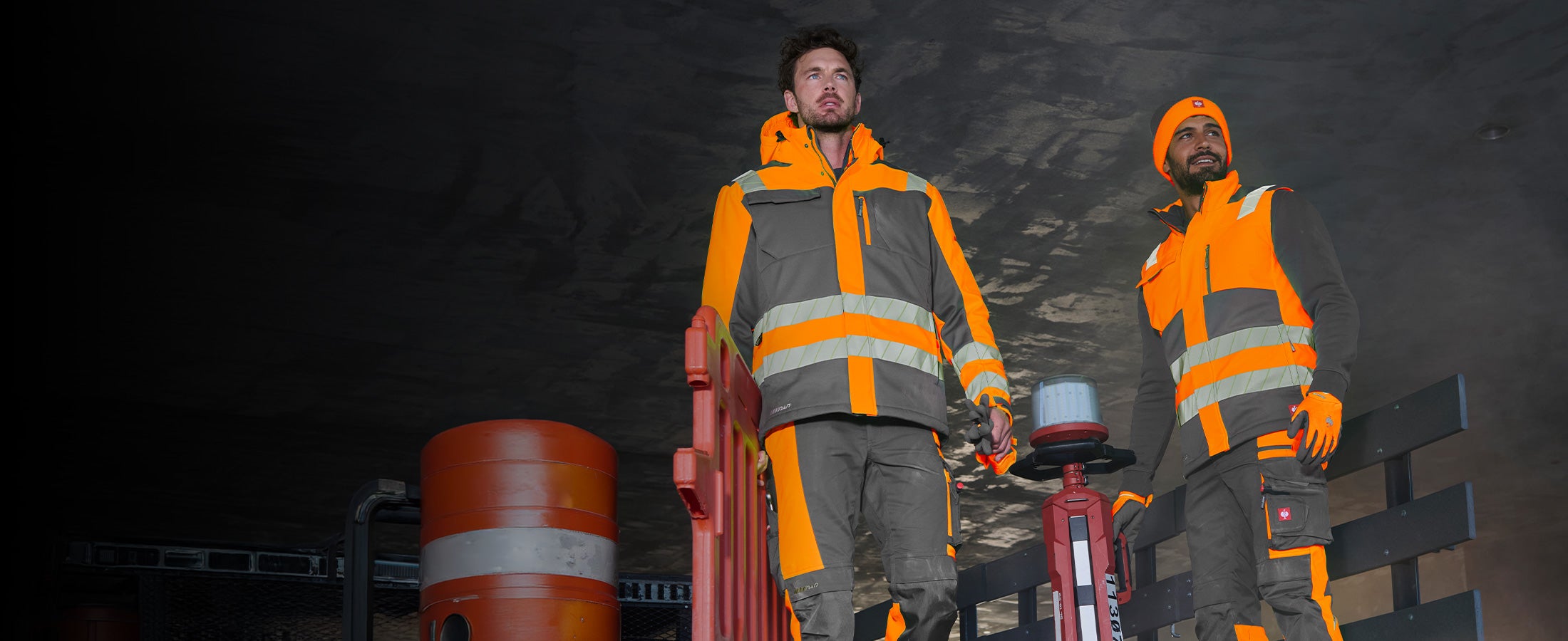 Zwei Männer in Warnschutzkleidung aus der motion24/7 Kollektion vor dunklem Bauwerk, einer davon mit Mütze.