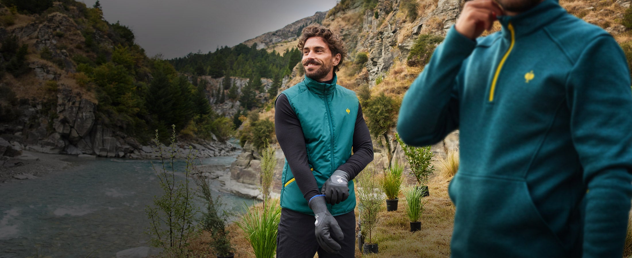 Zwei Männer in smaragdgrünem Troyer und Weste der e.s.trail Kollektion an einem Fluss in bergiger Landschaft.