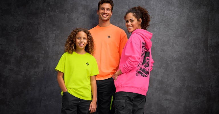 Een man in een neonoranje vintage shirt, een vrouw in een neonpink vintage hoodie en een kind in een neongeel vintage shirt staan samen voor een grijze betonnen muur.