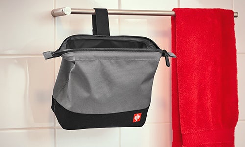 Une trousse de toilette grise avec un fond noir est suspendue à une barre métallique devant un mur carrelé blanc. La trousse est fixée à la barre par une boucle noire et arbore un petit logo rouge de la marque sur le devant. Une serviette rouge est suspendue à côté sur la même barre.