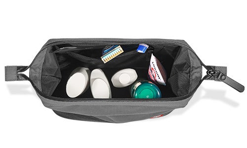 Trousse de toilette grise ouverte (washbag) avec des poignées latérales et un petit logo “e.s.”, remplie de plusieurs contenants de voyage blancs, d’une brosse à dents, d’un tube de dentifrice bleu et d’un petit pot vert.