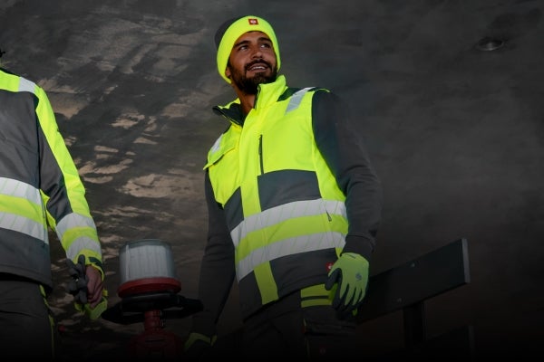 Deux hommes en vêtements haute visibilité de la collection motion24/7 devant une structure sombre, dont l’un porte un bonnet.