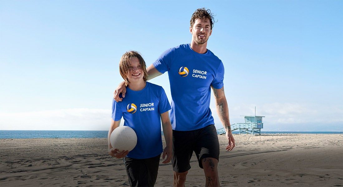 Een man in een blauw e.s. T-shirt met de opdruk Senior Kapitän en een jongen in een blauw e.s. T-shirt met de opdruk Junior Kapitän lopen op het strand met een volleybal
