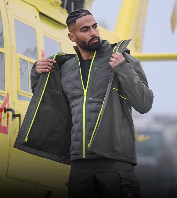 Mann in grauer Arbeitsjacke mit neongelben Reißverschlüssen vor einem gelben Hubschrauber.