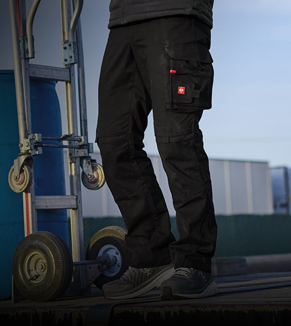 Schwarze Arbeitshose mit Seitentaschen an den Beinen, getragen mit grauen Schuhen neben einer Sackkarre.