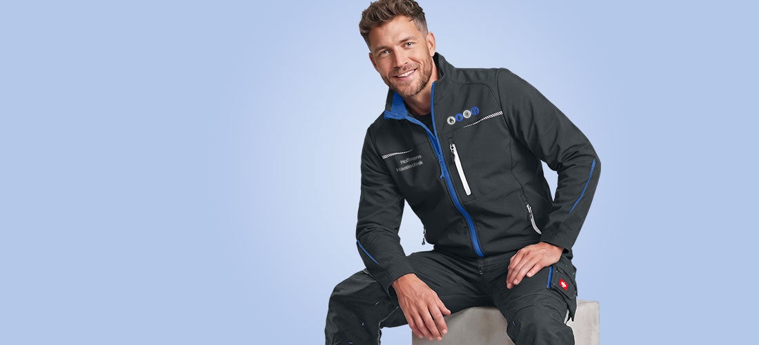 Sitzender Mann in schwarzer Arbeitskleidung mit blauem Besatz und Stickmotiv auf der Jacke vor hellblauem Hintergrund.