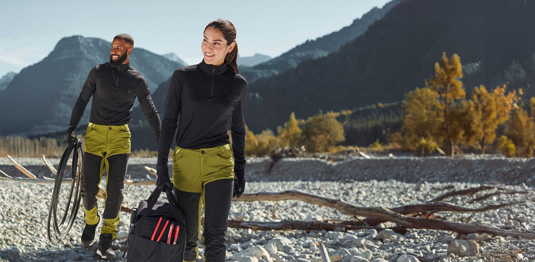 La femme et l’homme portent un pantalon de fonction e.s.trail en vert de genièvre / vert citron et un pull camionneur en noir
