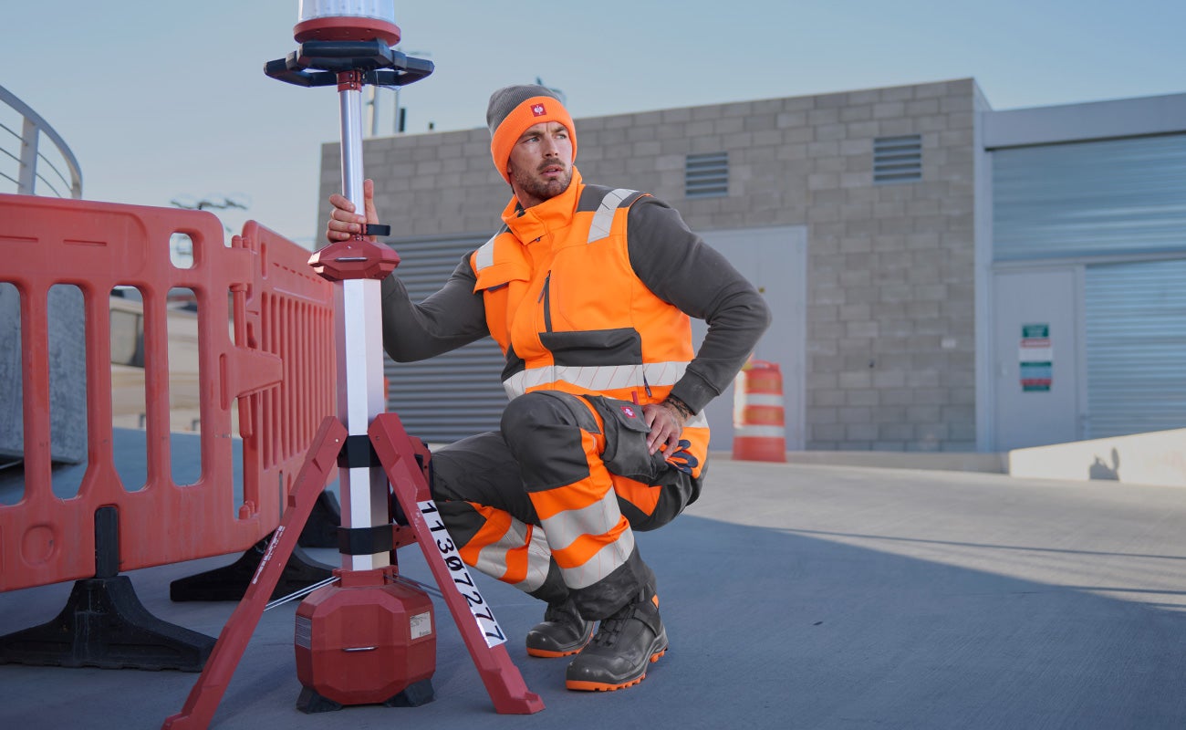 Motion 24/7 day and night: kneeling man in orange high-visibility workwear next to a site light and barrier.