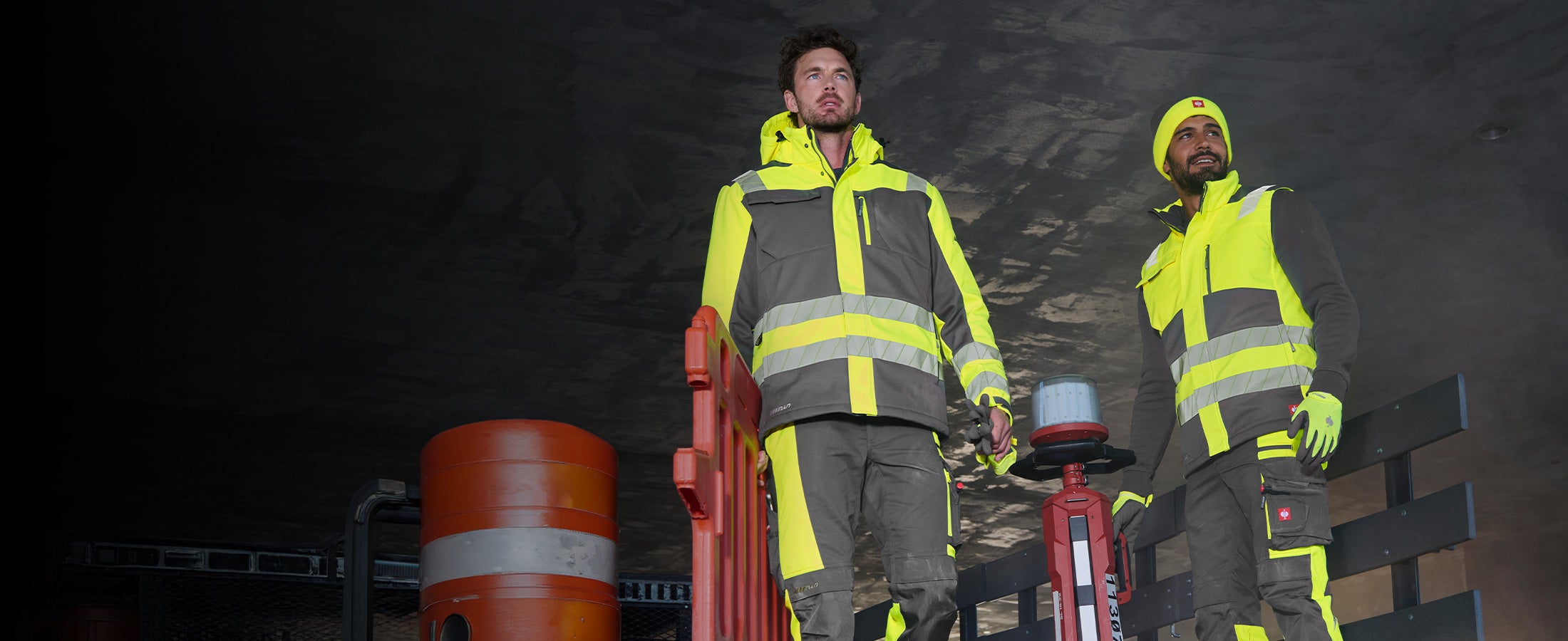 Two men in high-visibility clothing from the motion24/7 collection in front of a dark structure, one of them wearing a beanie.