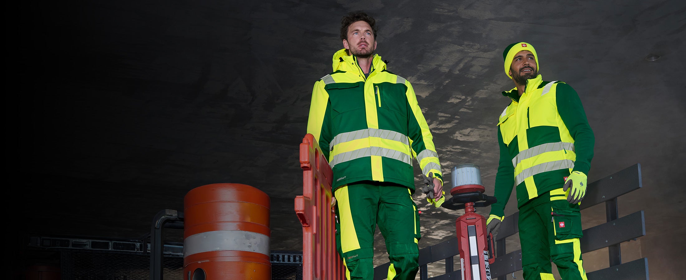 Two men in high-visibility clothing from the motion24/7 collection in front of a dark structure, one of them wearing a beanie.
