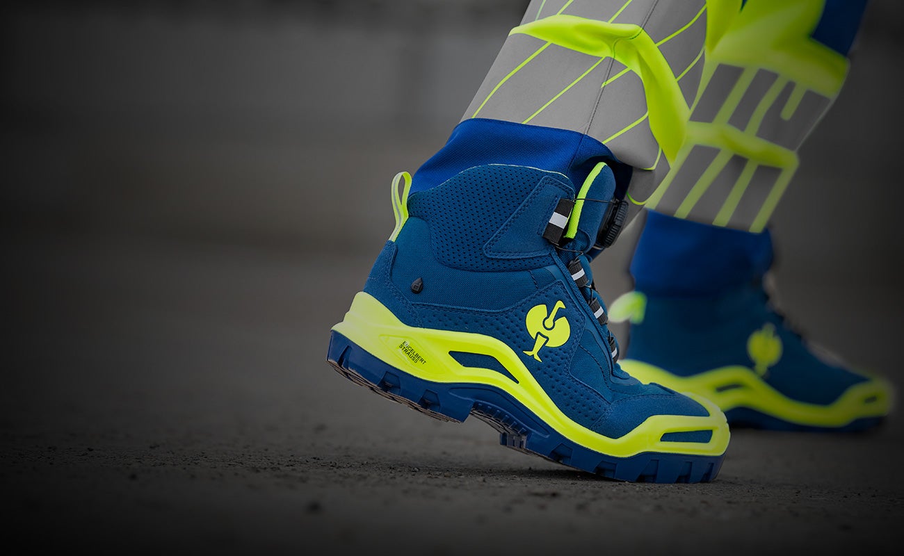 Blue S3 safety shoe e.s. Kastra II mid on a leg wearing grey high-visibility trousers with yellow details.