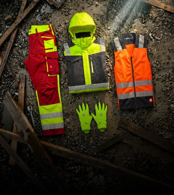 AI-generated / modified - Items from the motion24/7 collection are laid out as a jacket, safety vest, trousers and gloves on rocky ground.