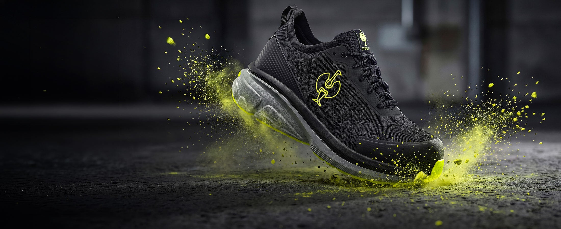 Chaussure de sécurité noire avec des accents jaunes devant des éclaboussures de peinture vert-jaune.