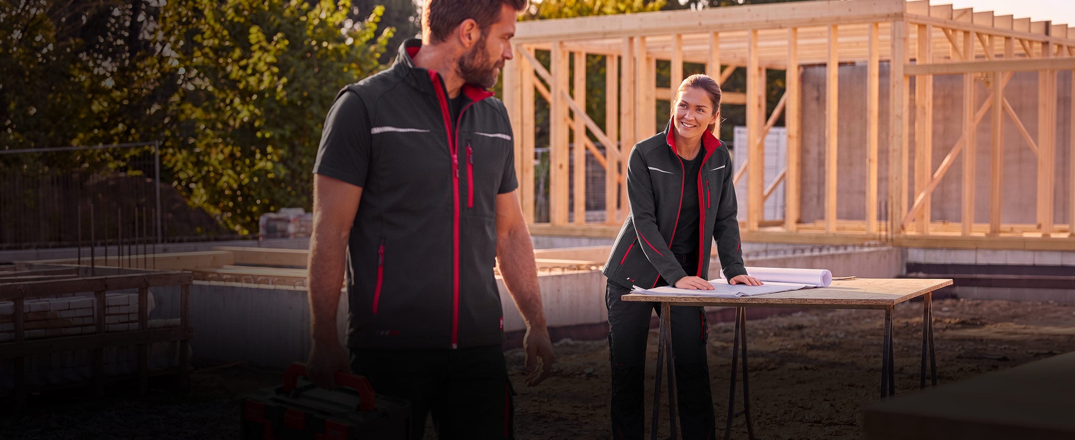Homme et femme en vêtements de travail graphite rouge feu devant une ossature en bois.