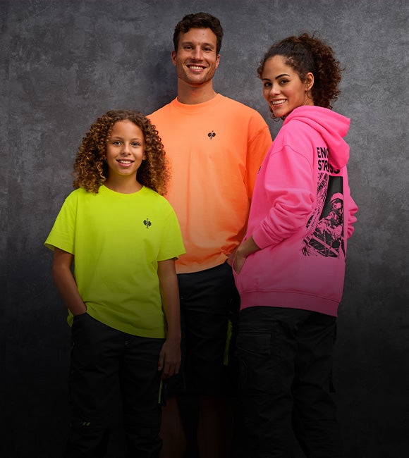 A man in a neon orange vintage shirt, a woman in a neonpink vintage hoody and a child in a neon yellow vintage shirt standing together in front of a grey concrete wall.