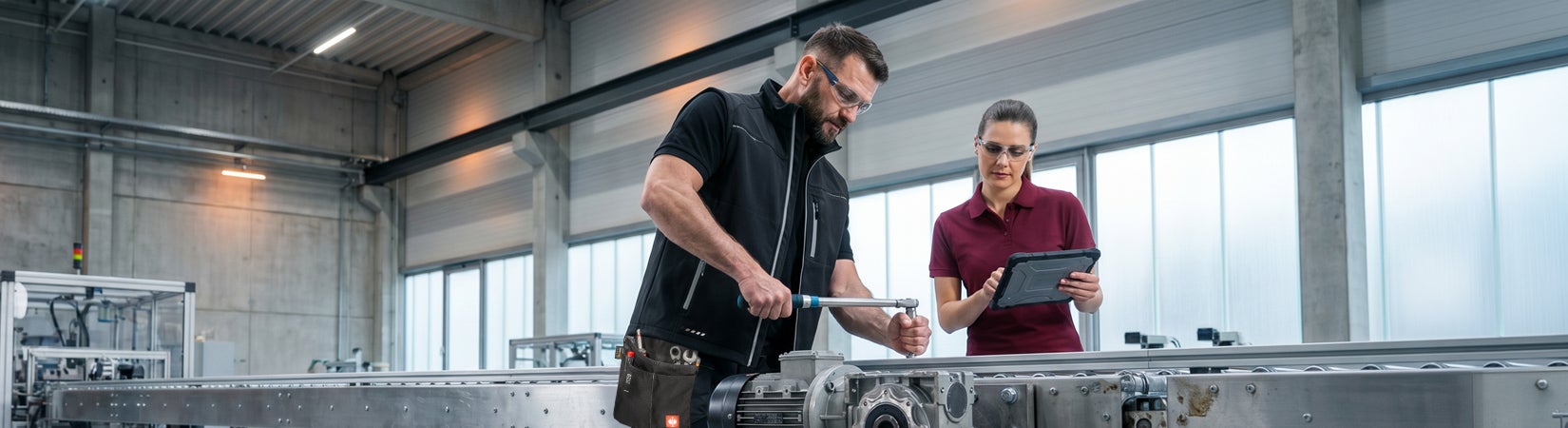 KI-generiert / modifiziert - Zwei Personen mit Schutzbrillen arbeiten an einer Metallanlage in einer hellen Werkhalle, eine davon mit Tablet.