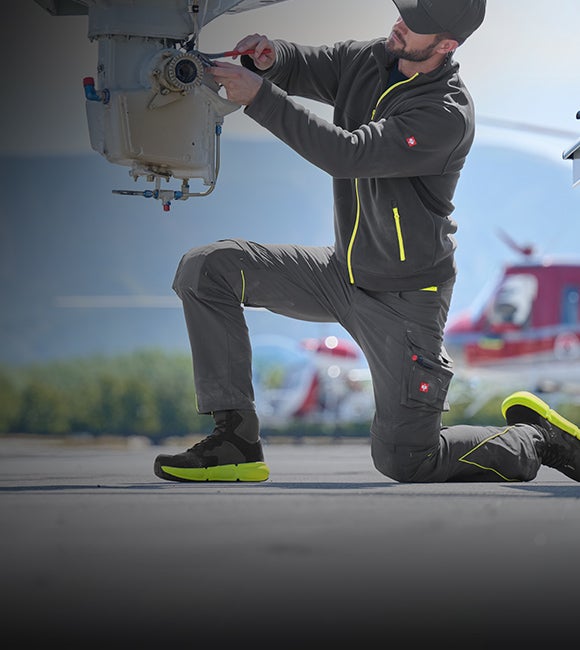 Kneeling man in dark workwear uses pliers on a helicopter.