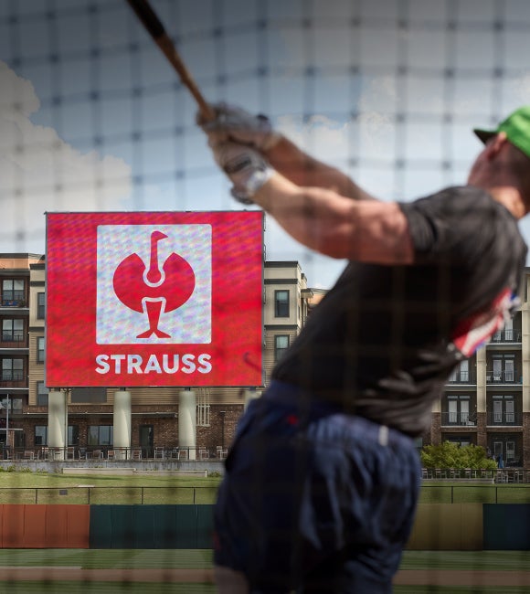 Unscharfer Baseballspieler vor einem großen STRAUSS-Schild hinter einem Netz.