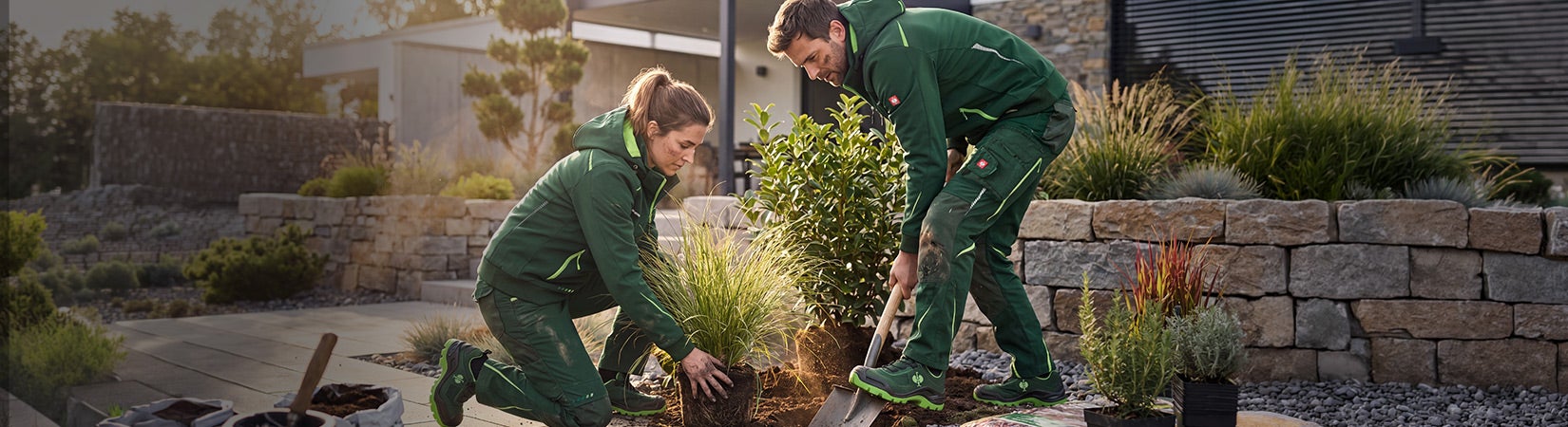 KI-generiert / modifiziert - Zwei Personen in grüner Arbeitskleidung pflanzen Sträucher in einem Garten.