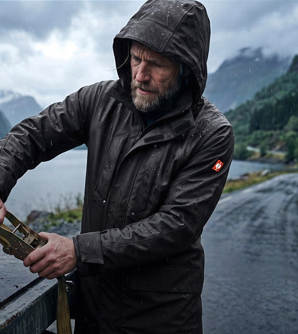 KI-generiert / modifiziert - Man in the Rain Parka e.s.line.nordic tightens a ratchet strap on a truck bed in the rain, with mountains in the background.