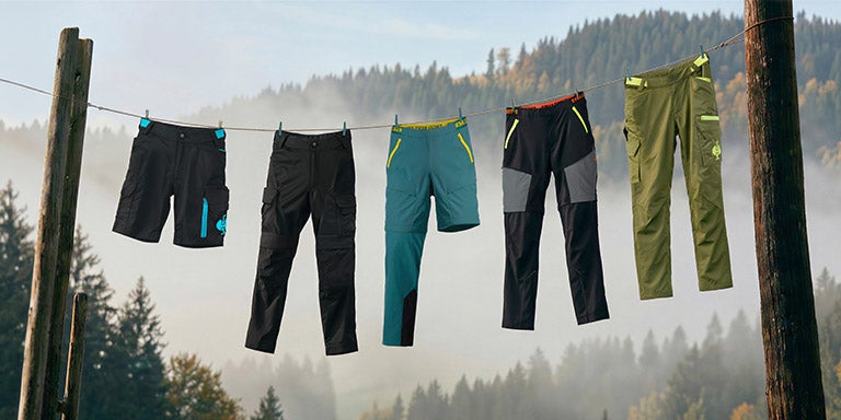 Cinq pantalons de travail sont suspendus à une corde devant une forêt brumeuse.