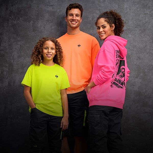 Un homme vêtu d'une t-shirt vintage orange néon, une femme vêtue d'un pull à capuche vintage rose néon et un enfant vêtu d'un t-shirt vintage jaune néon se tiennent ensemble devant un mur de béton gris.