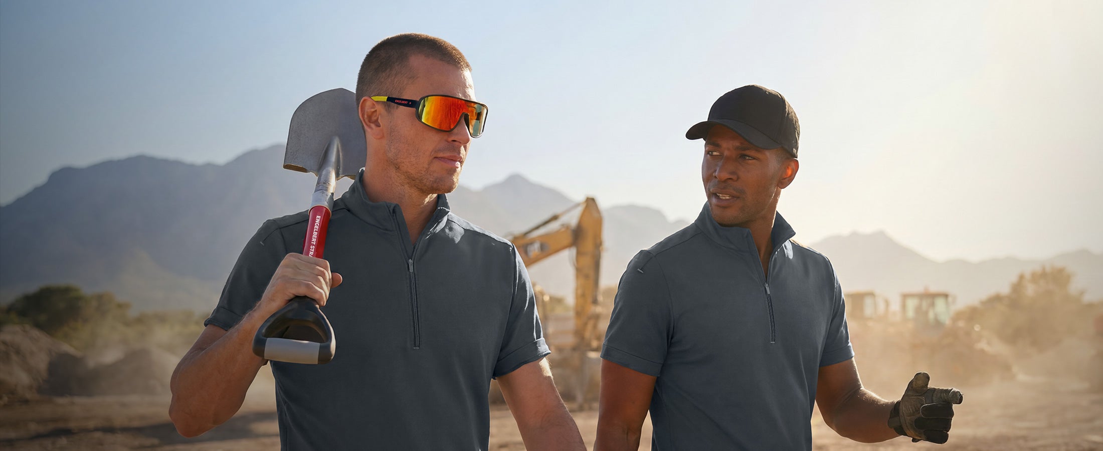 AI-generated / modified - Two men in blue work tops stand on a dusty construction site, one carrying a shovel on his shoulder.