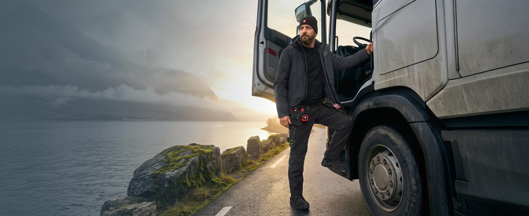 KI-generiert / modifiziert - Man i Vinterparkas e.s.line.nordic och Stretch hölstermidjebyxa e.s.line.nordic kliver ur en lastbil på en kustväg.
