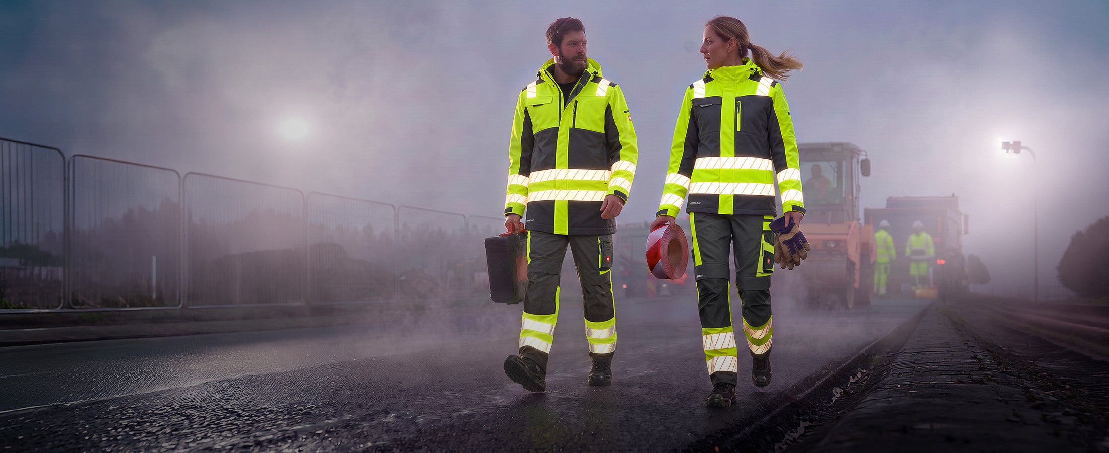 AI-genererad / modifierad: En man och en kvinna i varselkläder från kollektionen e.s.ambition går över en dimmig vägarbetsplats.