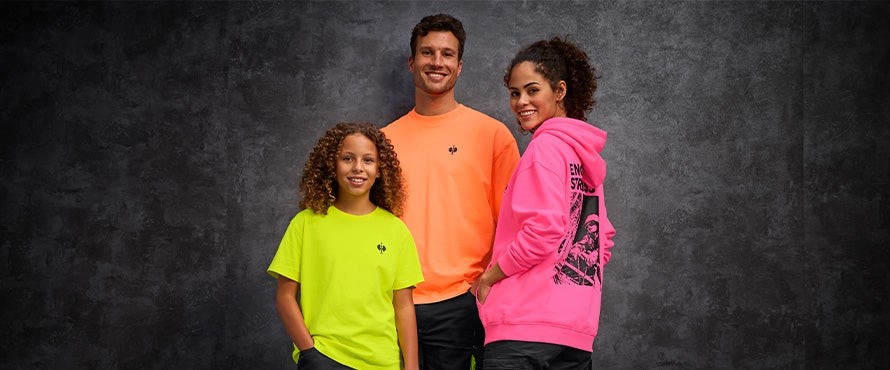 A man in a neon orange vintage shirt, a woman in a neonpink vintage hoody and a child in a neon yellow vintage shirt standing together in front of a grey concrete wall.
