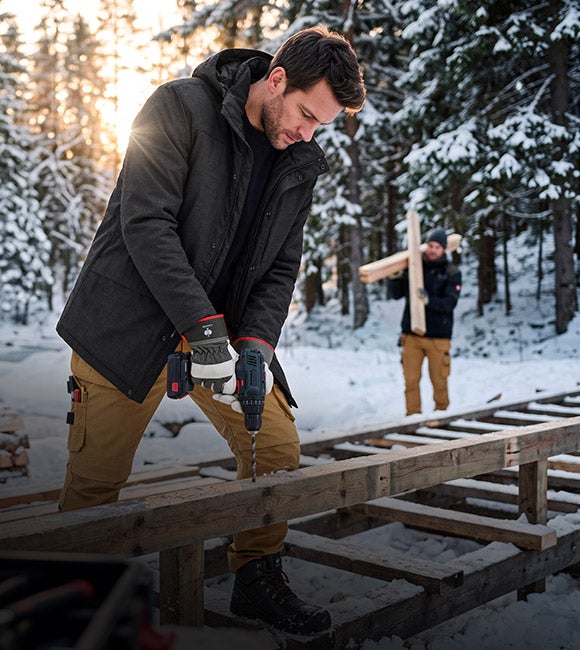 KI-generiert / modifiziert - Man i Vinterparkas e.s.line.nordic och Hölster-byxa e.s.line.nordic canvas borrar i en träbalk på en snötäckt byggarbetsplats.